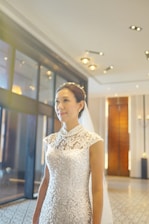 A woman wearing a detailed lace wedding dress with a veil standing indoors in a well-lit area. The hallway features elegant decor with a patterned floor, ambient lighting, and large windows.