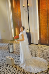 A bride in an elegant white wedding dress stands on a patterned floor, holding a bouquet of red flowers. The background features tall wooden panels and a standing candle holder, creating a warm, sophisticated atmosphere. A small suitcase is seen on the floor beside her.