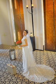A bride in an elegant white wedding dress stands on a patterned floor, holding a bouquet of red flowers. The background features tall wooden panels and a standing candle holder, creating a warm, sophisticated atmosphere. A small suitcase is seen on the floor beside her.