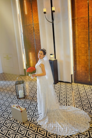 A bride in an elegant white wedding dress stands on a patterned floor, holding a bouquet of red flowers. The background features tall wooden panels and a standing candle holder, creating a warm, sophisticated atmosphere. A small suitcase is seen on the floor beside her.