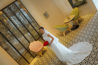 A bride in a white lace wedding gown stands holding a bouquet of red roses. The background features intricate glass paneling, a geometric patterned floor, and a seating area with two green chairs and a small round table.