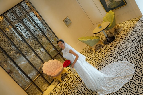 A bride in a white lace wedding gown stands holding a bouquet of red roses. The background features intricate glass paneling, a geometric patterned floor, and a seating area with two green chairs and a small round table.