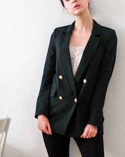 A person stands against a plain background, wearing a dark green blazer with gold buttons over a white lace top and black pants. The person's posture is relaxed, with one hand slightly bent and resting by the side.