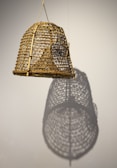 A woven bamboo or rattan lampshade hangs against a plain background. The intricate lattice work is highlighted by light, casting a distinct shadow on the wall that mirrors its pattern.