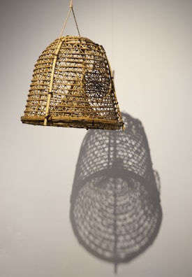 A woven bamboo or rattan lampshade hangs against a plain background. The intricate lattice work is highlighted by light, casting a distinct shadow on the wall that mirrors its pattern.