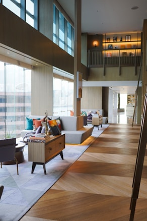 The image depicts a modern interior space with large windows allowing ample natural light. There are couches with colorful cushions, side tables, bookshelves, and various décor items. The space features a two-level structure with wooden floors and contemporary furniture. Shelves on the upper level showcase books and decorative objects.