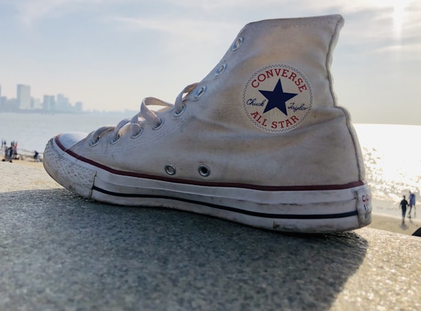 A well-worn white Converse All Star sneaker rests on a concrete surface with the sun reflecting off a large body of water in the background. The silhouette of a distant city skyline is visible, along with a few small figures walking at the water's edge.