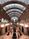 An architectural interior featuring a grand arched ceiling with intricate patterns and numerous windows, casting soft light over a spacious hall. At the center is a sculpture of figures holding a large spherical object. People are walking or sitting along the sides, engaging with the surrounding artwork and exhibits.