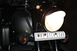 A close-up view of a motorcycle features prominently, highlighting the bright headlight and the license plate with the number PY 01 CV 2046. The vehicle has a dark, matte finish with subtle reflections indicating a sleek design. The surroundings are dimly lit, emphasizing the light sources on the motorcycle.