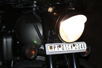 A close-up view of a motorcycle features prominently, highlighting the bright headlight and the license plate with the number PY 01 CV 2046. The vehicle has a dark, matte finish with subtle reflections indicating a sleek design. The surroundings are dimly lit, emphasizing the light sources on the motorcycle.
