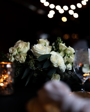 A romantic setting featuring an arrangement of white roses surrounded by green foliage. Soft, ambient lighting from bokeh lights in the background creates a warm and intimate atmosphere. A reflective wine glass and a decorative candle holder are also present, adding to the elegant setting.