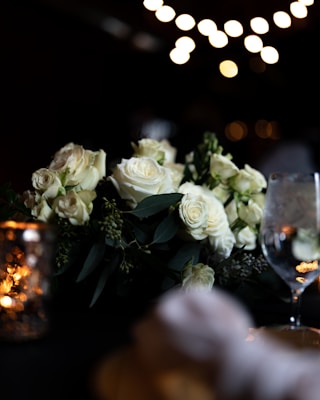 A romantic setting featuring an arrangement of white roses surrounded by green foliage. Soft, ambient lighting from bokeh lights in the background creates a warm and intimate atmosphere. A reflective wine glass and a decorative candle holder are also present, adding to the elegant setting.