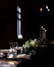 A dimly lit dining table is elegantly arranged with a centerpiece of white and green flowers. The table is set with crystal clear glasses, folded napkins, and ornate silver candle holders, creating a sophisticated and intimate atmosphere.