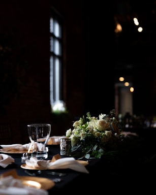 A dimly lit dining table is elegantly arranged with a centerpiece of white and green flowers. The table is set with crystal clear glasses, folded napkins, and ornate silver candle holders, creating a sophisticated and intimate atmosphere.