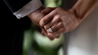 Two hands are clasped together, symbolizing a connection or relationship. One hand is adorned with a prominent diamond ring, likely indicating engagement or marriage. The background is softly blurred, emphasizing the close-up of the hands.