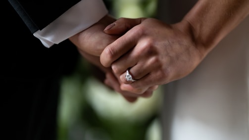 Two hands are clasped together, symbolizing a connection or relationship. One hand is adorned with a prominent diamond ring, likely indicating engagement or marriage. The background is softly blurred, emphasizing the close-up of the hands.