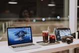 A workspace setup on a table by a window includes a laptop with an island wallpaper, a tablet displaying photo thumbnails, a camera, and two glasses filled with iced beverages. The background shows a blurred reflection of a person.