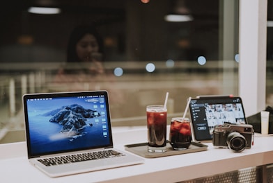 A workspace setup on a table by a window includes a laptop with an island wallpaper, a tablet displaying photo thumbnails, a camera, and two glasses filled with iced beverages. The background shows a blurred reflection of a person.