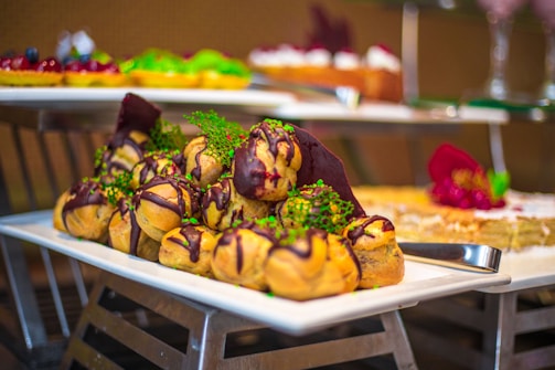A beautifully arranged tray of pastries, likely profiteroles or cream puffs, drizzled with chocolate and garnished with herbs and edible flowers. The tray is placed on a metal stand, and additional desserts can be seen blurred in the background, giving a sense of a dessert buffet.