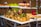 A beautifully arranged tray of pastries, likely profiteroles or cream puffs, drizzled with chocolate and garnished with herbs and edible flowers. The tray is placed on a metal stand, and additional desserts can be seen blurred in the background, giving a sense of a dessert buffet.
