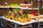 A beautifully arranged tray of pastries, likely profiteroles or cream puffs, drizzled with chocolate and garnished with herbs and edible flowers. The tray is placed on a metal stand, and additional desserts can be seen blurred in the background, giving a sense of a dessert buffet.