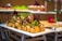 A beautifully arranged tray of pastries, likely profiteroles or cream puffs, drizzled with chocolate and garnished with herbs and edible flowers. The tray is placed on a metal stand, and additional desserts can be seen blurred in the background, giving a sense of a dessert buffet.