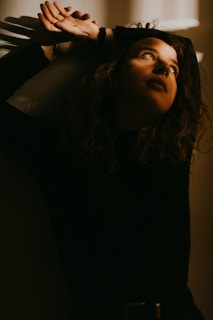 Light casts dramatic shadows across a person's face, highlighting their upward gaze and creating an atmospheric scene. The setting is dimly lit, with a warm tone emphasizing introspection and contemplation.