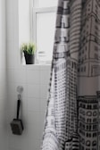 A bathroom interior featuring white tiled walls. A black and white patterned shower curtain partially covers the view. A black potted plant and a glass are placed on the window ledge. A brush with a long handle is hanging from a hook on the wall.