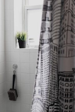 A bathroom interior featuring white tiled walls. A black and white patterned shower curtain partially covers the view. A black potted plant and a glass are placed on the window ledge. A brush with a long handle is hanging from a hook on the wall.