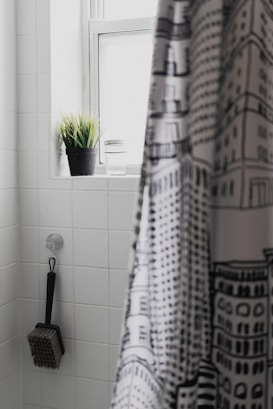 A bathroom interior featuring white tiled walls. A black and white patterned shower curtain partially covers the view. A black potted plant and a glass are placed on the window ledge. A brush with a long handle is hanging from a hook on the wall.