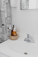 A bathroom scene with a white sink and a chrome faucet. There's a woven basket holding two soap dispensers on the sink. The wall behind the sink is covered with white tiles, and there's a mirror on the wall reflecting part of the room. A shower curtain with a black and white cityscape design is partially visible.