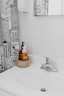 A bathroom scene with a white sink and a chrome faucet. There's a woven basket holding two soap dispensers on the sink. The wall behind the sink is covered with white tiles, and there's a mirror on the wall reflecting part of the room. A shower curtain with a black and white cityscape design is partially visible.