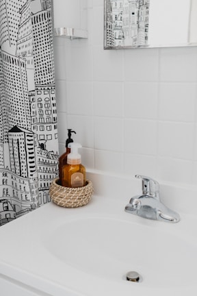 A bathroom scene with a white sink and a chrome faucet. There's a woven basket holding two soap dispensers on the sink. The wall behind the sink is covered with white tiles, and there's a mirror on the wall reflecting part of the room. A shower curtain with a black and white cityscape design is partially visible.