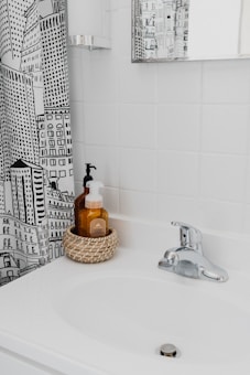 A bathroom scene with a white sink and a chrome faucet. There's a woven basket holding two soap dispensers on the sink. The wall behind the sink is covered with white tiles, and there's a mirror on the wall reflecting part of the room. A shower curtain with a black and white cityscape design is partially visible.