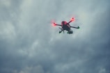 A drone with glowing green and red lights is flying against a backdrop of cloudy, overcast skies.