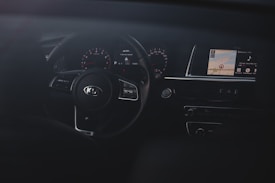 The interior of a car focusing on the dashboard and steering wheel. The steering wheel has a Kia logo and is surrounded by gauges that show speed and other metrics. A digital display screen is visible on the right showing map directions and multimedia information.