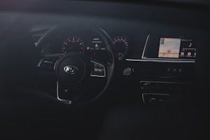 The interior of a car focusing on the dashboard and steering wheel. The steering wheel has a Kia logo and is surrounded by gauges that show speed and other metrics. A digital display screen is visible on the right showing map directions and multimedia information.