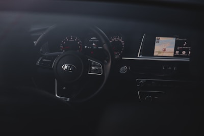 The interior of a car focusing on the dashboard and steering wheel. The steering wheel has a Kia logo and is surrounded by gauges that show speed and other metrics. A digital display screen is visible on the right showing map directions and multimedia information.