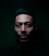 A man's face is partially illuminated against a dark background, creating a dramatic shadow effect. The lighting highlights his facial features, particularly the structure of his cheekbones and lips. His hair is styled neatly, and he maintains a neutral expression.