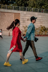 A couple is walking hand in hand. The woman is wearing a bright red dress and yellow shoes, holding a water bottle. The man is in a denim jacket, dark pants, red-tinted shoes, and a black cap, carrying a camera. They are outdoors, with a stone wall in the background adorned with detailed carvings, nearby foliage, and a few other people in the distance.