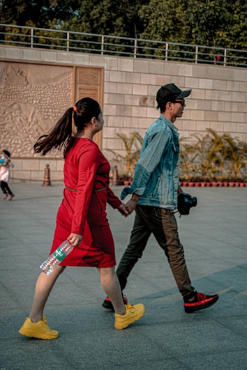 A couple is walking hand in hand. The woman is wearing a bright red dress and yellow shoes, holding a water bottle. The man is in a denim jacket, dark pants, red-tinted shoes, and a black cap, carrying a camera. They are outdoors, with a stone wall in the background adorned with detailed carvings, nearby foliage, and a few other people in the distance.