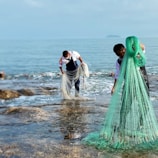 Two people are standing near the water, handling large fishing nets. They are wearing waterproof overalls and appear to be collecting or organizing the nets along a rocky shore. The sea is calm and the skies are clear, suggesting a peaceful and industrious setting.