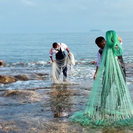 Two people are standing near the water, handling large fishing nets. They are wearing waterproof overalls and appear to be collecting or organizing the nets along a rocky shore. The sea is calm and the skies are clear, suggesting a peaceful and industrious setting.