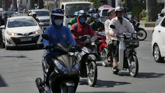 A busy urban street features multiple individuals riding scooters. They are wearing helmets and masks, suggestive of traffic safety and health precautions. A taxi and other vehicles are visible in the background, indicating a bustling city environment.