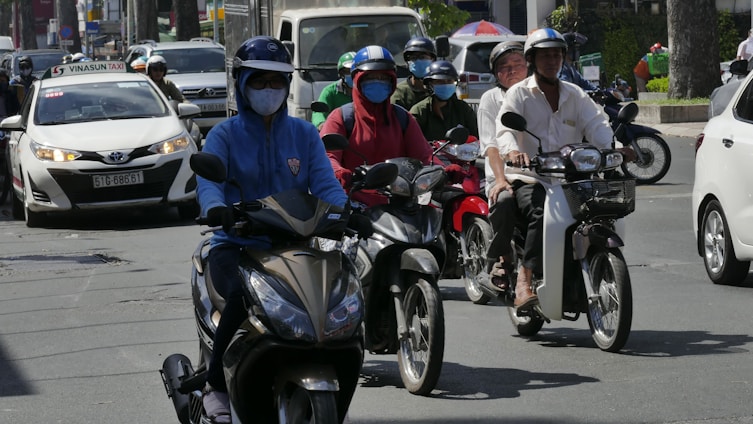 A busy urban street features multiple individuals riding scooters. They are wearing helmets and masks, suggestive of traffic safety and health precautions. A taxi and other vehicles are visible in the background, indicating a bustling city environment.