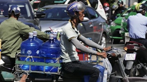 A man wearing a helmet rides a motorcycle through a busy traffic area. He is transporting several large blue water bottles secured on the rear of the bike. Other motorcycles and cars are visible in the background.