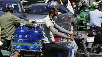 A man wearing a helmet rides a motorcycle through a busy traffic area. He is transporting several large blue water bottles secured on the rear of the bike. Other motorcycles and cars are visible in the background.