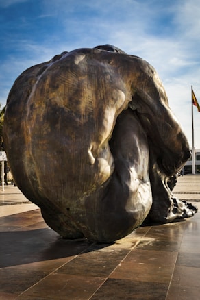 A massive bronze sculpture depicting a person crouching with their head tucked between their knees, creating a rounded, hunched posture. The texture of the sculpture's surface features smooth and rough patches, enhancing its three-dimensional quality.