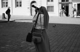 A person wearing a headscarf and long skirt is holding a professional camera to their eye, aiming it at something beyond the frame. They are standing on a cobblestone street, and in the background, there are buildings with tall windows. A couple of other people are visible in the distance, along with a pigeon on the ground.