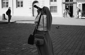 A person wearing a headscarf and long skirt is holding a professional camera to their eye, aiming it at something beyond the frame. They are standing on a cobblestone street, and in the background, there are buildings with tall windows. A couple of other people are visible in the distance, along with a pigeon on the ground.